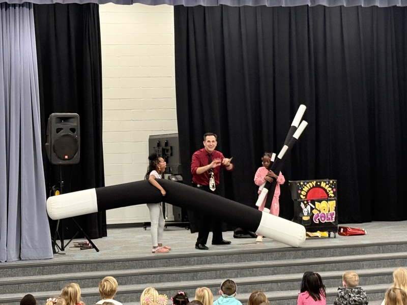 Magician on stage performing for students sitting and watching in the commons.  Students are holding blow up wands.