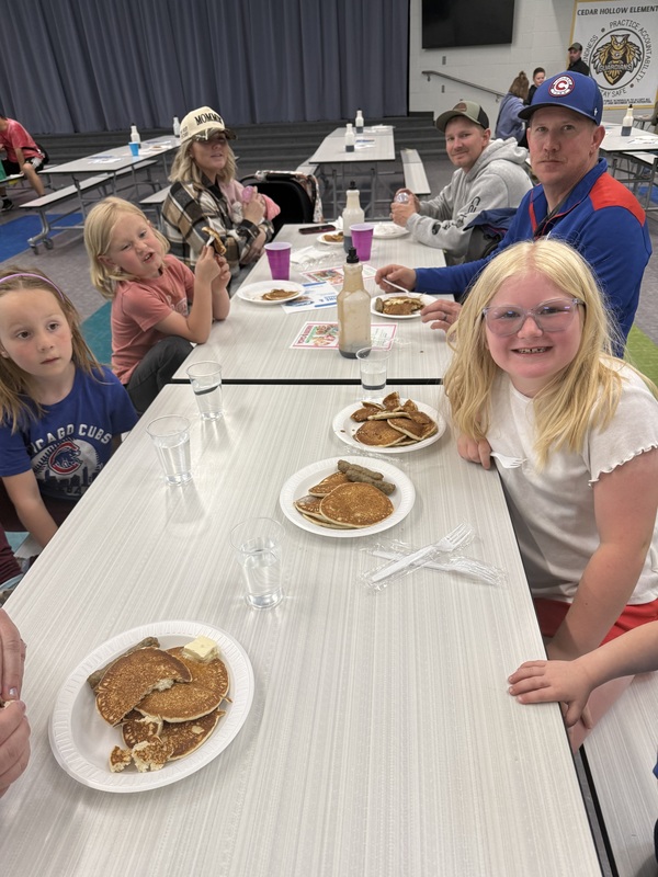 Students seated at lunch tables eating pancakes. 3 girls smiling with their family.