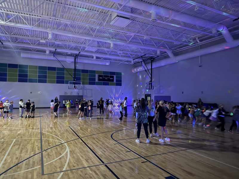 Students standing and dancing in the gym with colorful lights on the ceiling.