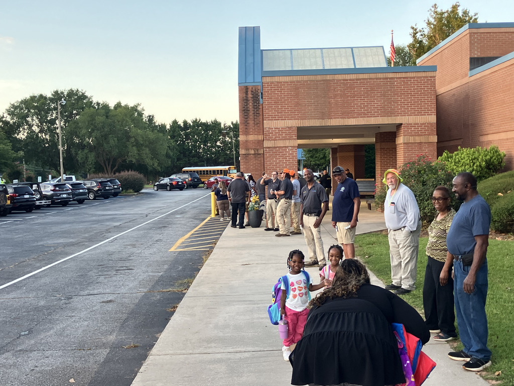 people walking up sidewalk with volunteers greeting towards front of school