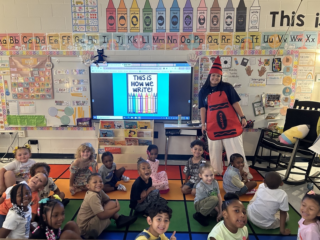 Teacher dressed up as a crayon with students on carpet and the board reading "This is How We Write"