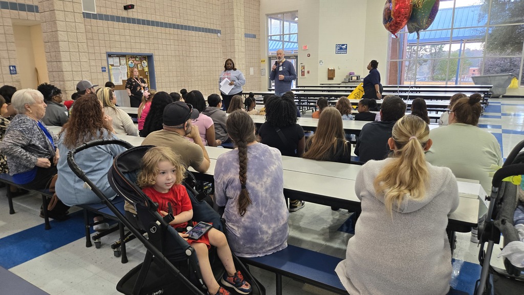 parents and students at tables in cafeteria, 2 adults in front talking