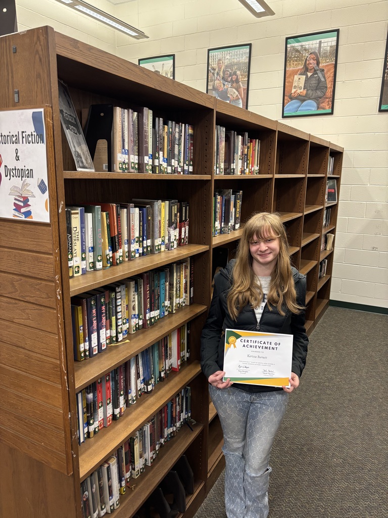 Student Karissa Burnett holds her certificate in front of the Historical Fiction section of the library.