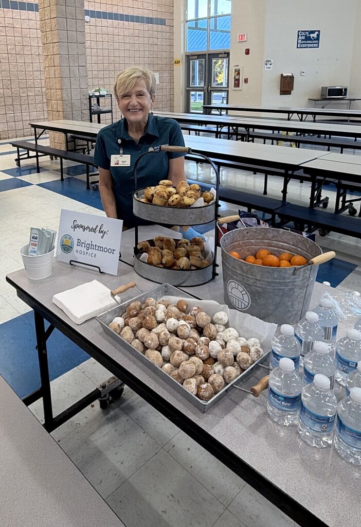 lady in front of table with muffins, donut holes, cuties, and water. Sign reads "Sponsored by Brightmoor Hospice"