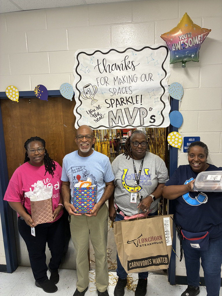 Four Cowan Road Elementary custodians pose together in front of a decorated doorway while holding gift bags and a LongHorn Steakhouse bag. Above them is a handmade sign that reads, “Thanks for making our spaces sparkle! You’re the real MVP’s.” Balloons and gold decorations surround the sign as the custodians smile for the photo.