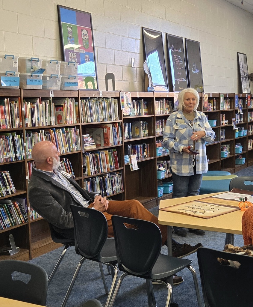 A woman speaking while standing in front of media center bookshelves next to a seated colleague.