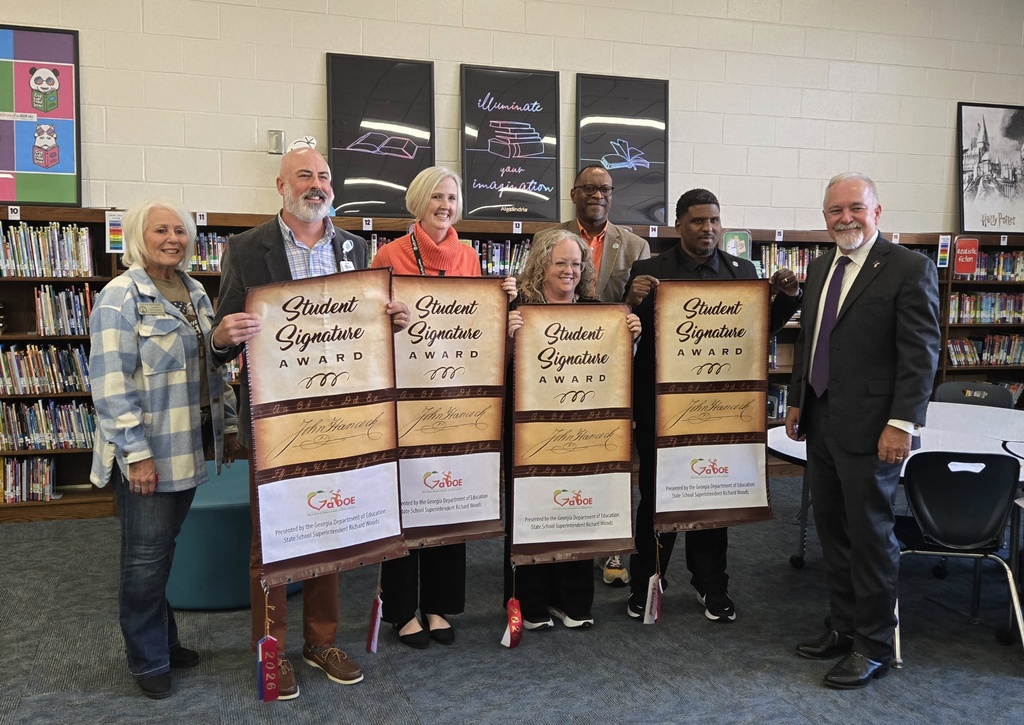 Seven educators and officials standing in a school library, holding four separate "Student Signature Award" banners.
