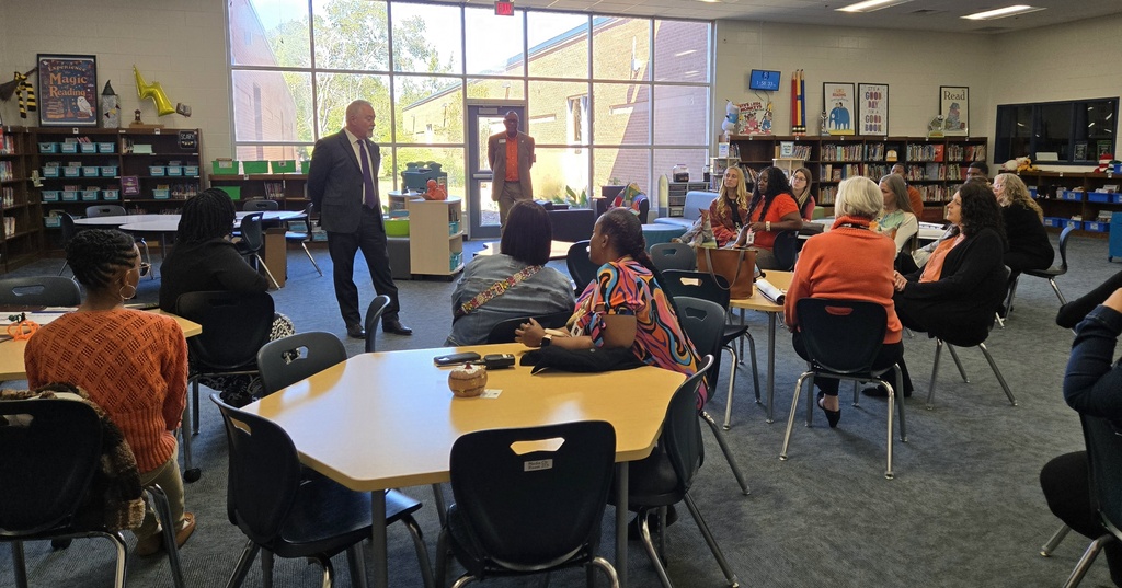 A speaker standing in a school library during a presentation to educators seated around tables.