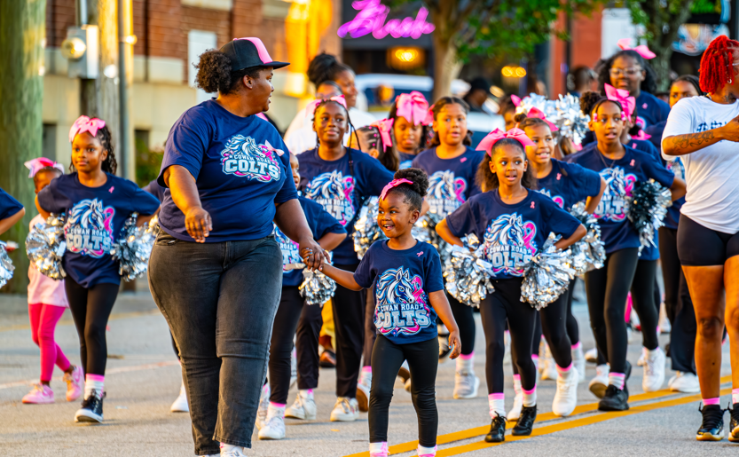 A small cheerleader walking hand-in-hand with an adult volunteer down the parade route followed by her team.