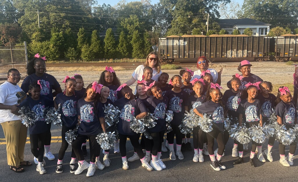 The entire Cowan Colt cheer team lined up in formation in a parking lot, holding silver pom-poms and wearing matching pink hair bows.