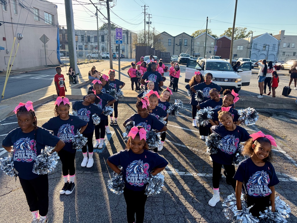 The entire Cowan Colt cheer team lined up in formation in a parking lot, holding silver pom-poms and wearing matching pink hair bows.