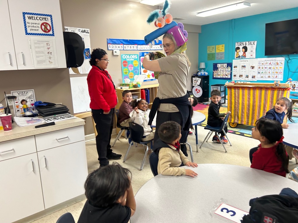 An adult is giving a puppet presentation with a colorful and funny puppet on their head in a Pre-K classroom