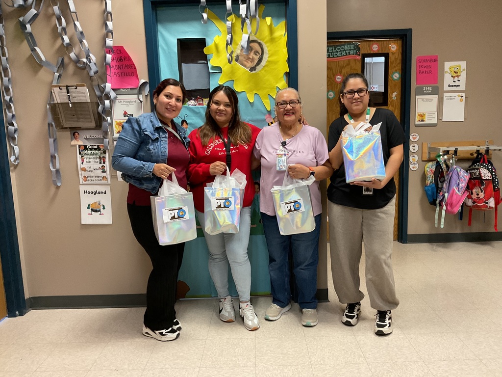 A group of ladies smiling to the camera with an appreciation gift outside a classroom.