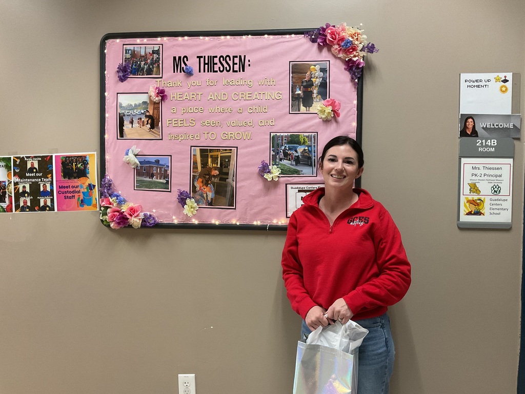 A female principal standing outside her office with an appreciation gift in front of a bulletin board with a gratitude message.