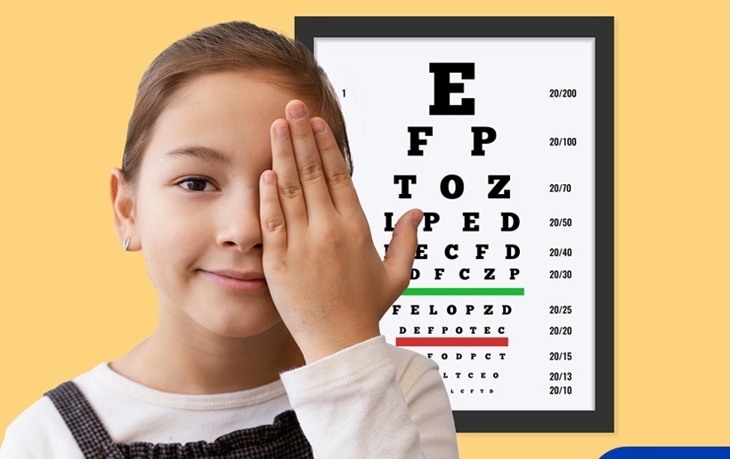 Student covering one eye while looking at camera with a vision test poster  behind her.  