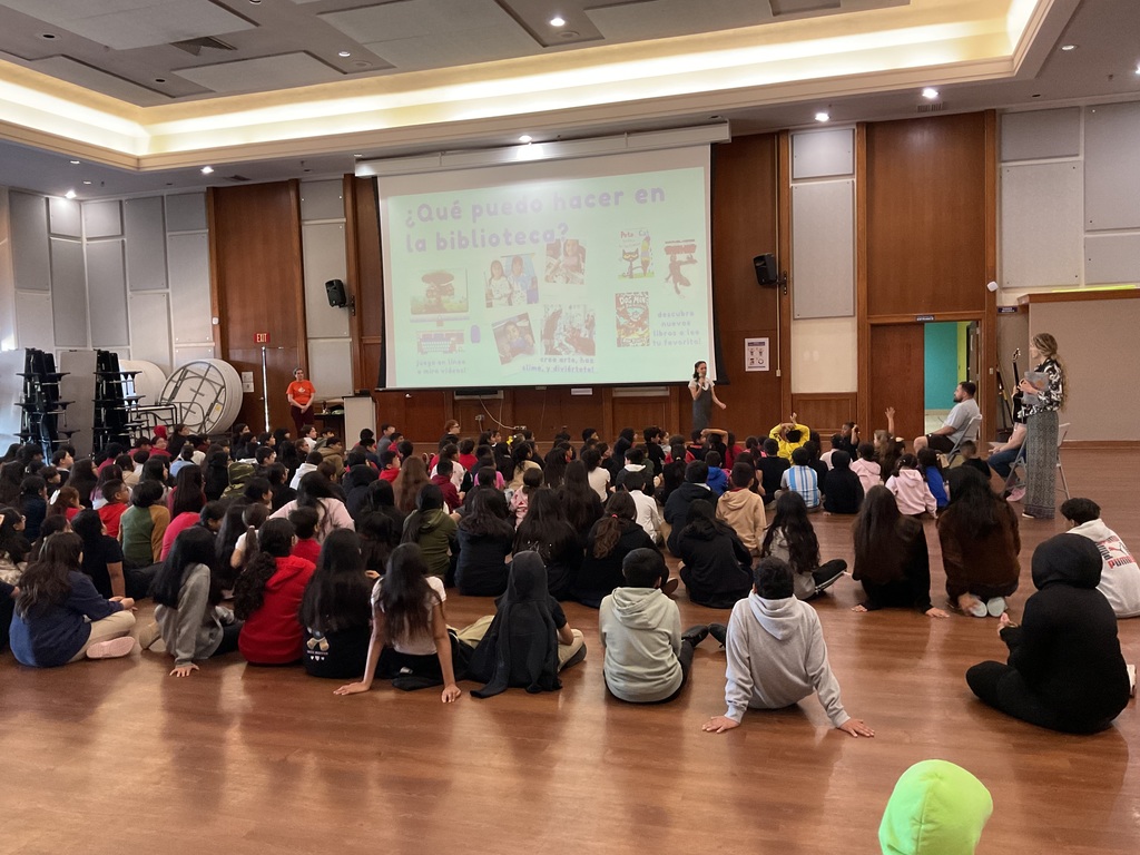 A group of students sitting on the floor looking a t big screen that says What can I do at the library? in Spanish