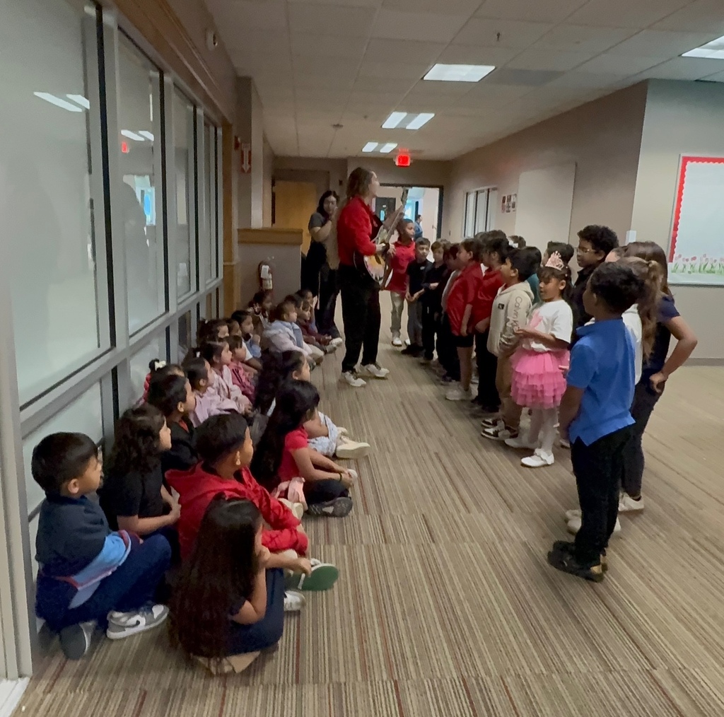 A group of students sitting on the floor while a group is standing up in front of them with a teacher and a guitat