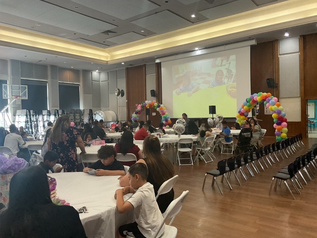 School gymnasium with tables and a screen, where some parents and children are waiting for their Pre-K student’s graduation ceremony.