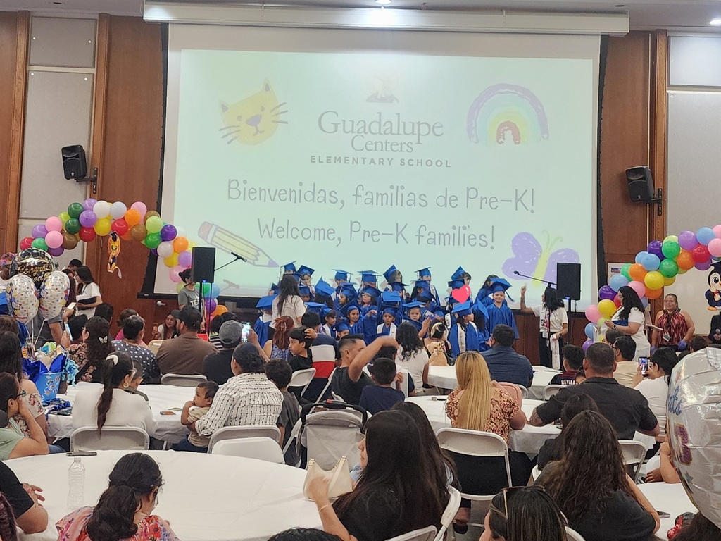 Graduation photograph of Pre-K children wearing blue caps and gowns, a group of 40 children while their parents watch them sing a song.
