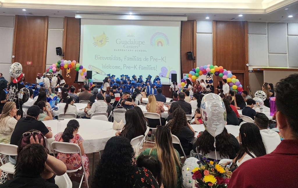 Graduation photograph of Pre-K children wearing blue caps and gowns, a group of 40 children while their parents watch them sing a song.