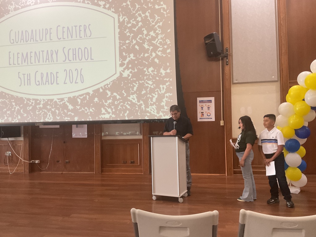 A man on a podium with a microphone and 2 students by his side