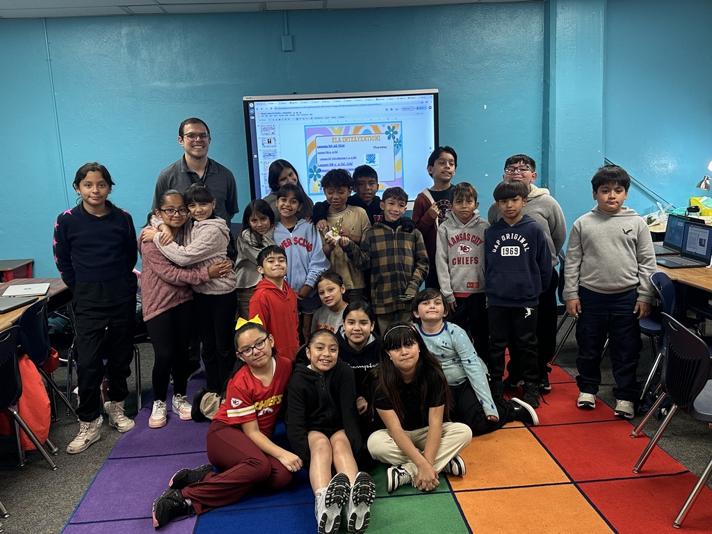 A group of student and teacher posing for a photo with a tv on the background and a colorful carpet.