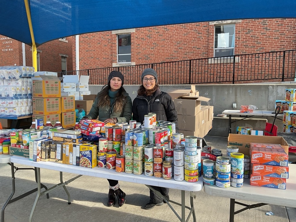Two volunteers holding cans of food and smiling at the camera while standing behind a table full of canned goods arranged for distribution.