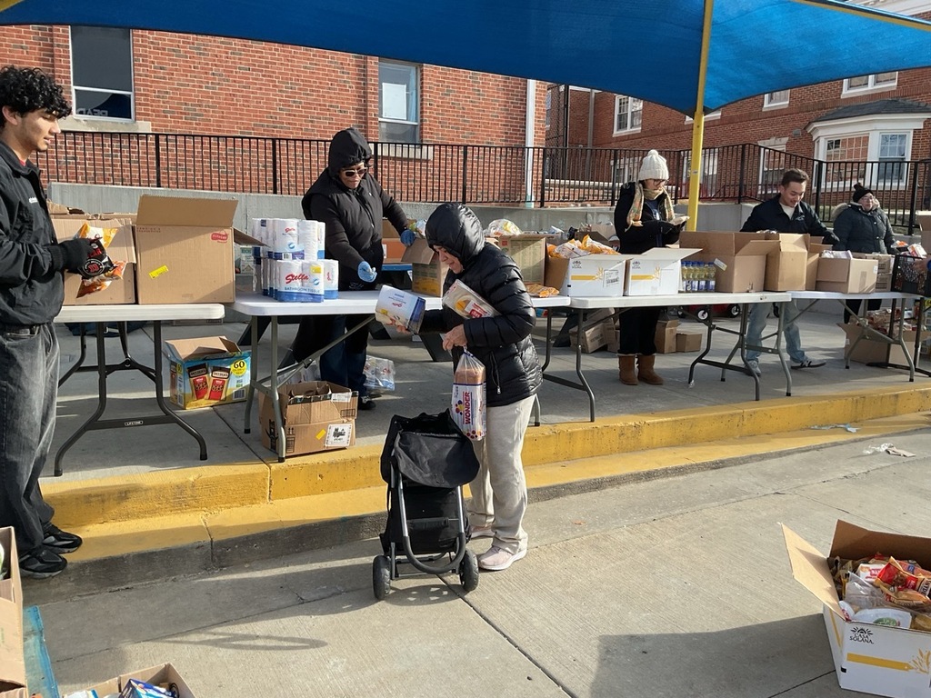 Volunteers are distributing food pantry items while people stand in line in an orderly manner to receive the products. The volunteers hand out food supplies as each person steps forward one by one, showing an organized and respectful food distribution process.