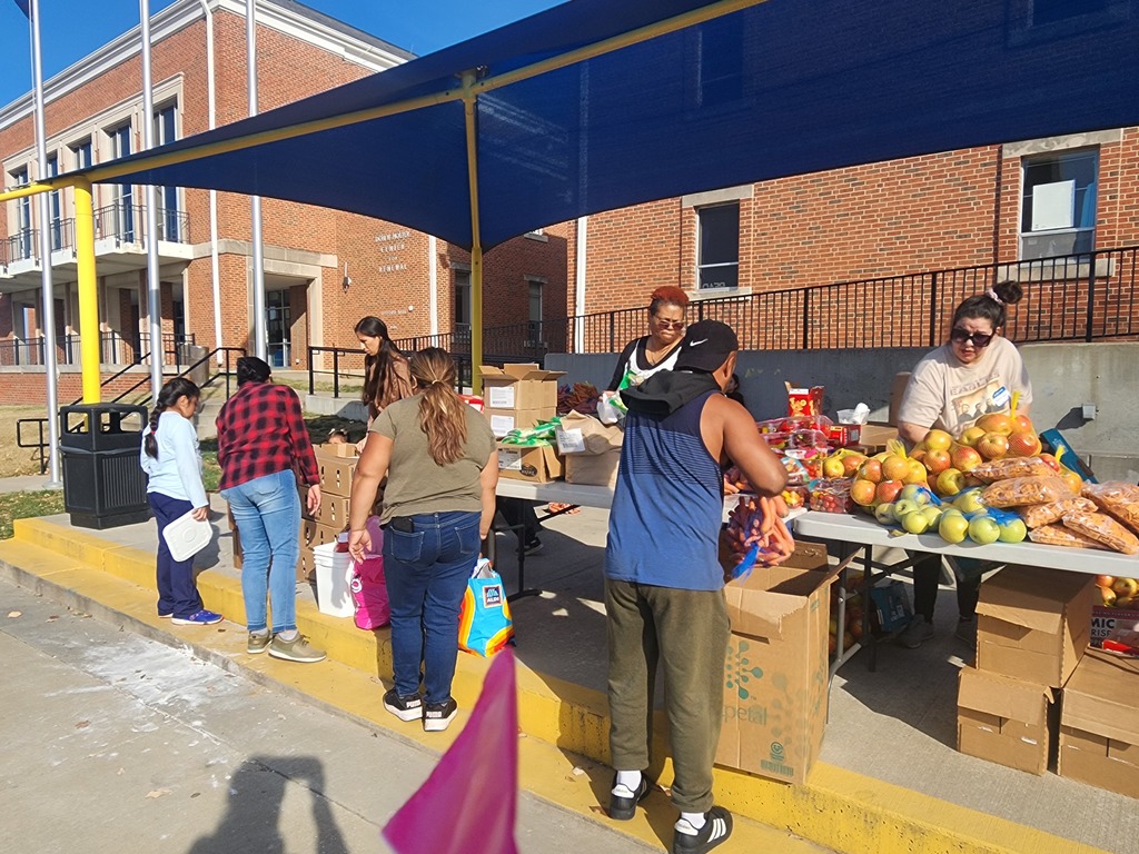 Volunteers are distributing food pantry items while people stand in line in an orderly manner to receive the products. The volunteers hand out food supplies as each person steps forward one by one, showing an organized and respectful food distribution process.