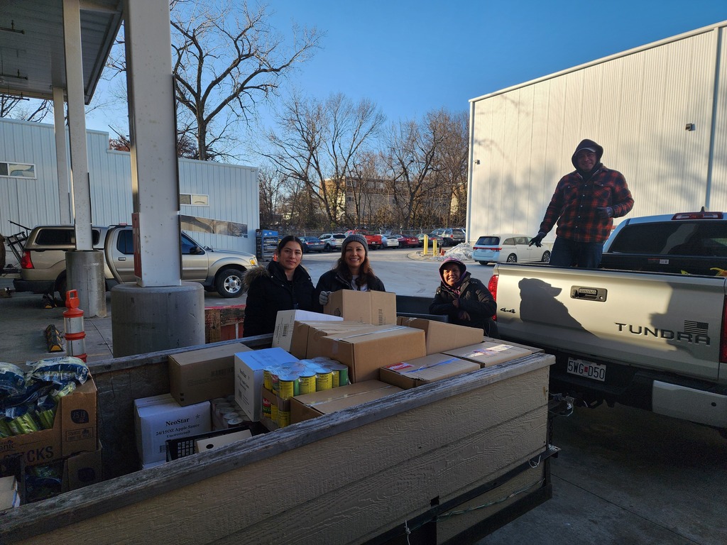 Volunteers are posing for a photo at a food donation site. They are at the location where food is collected and prepared to be taken to the school. The donated food will later be distributed to the families of students. The scene shows a group moment recognizing their support and community service.