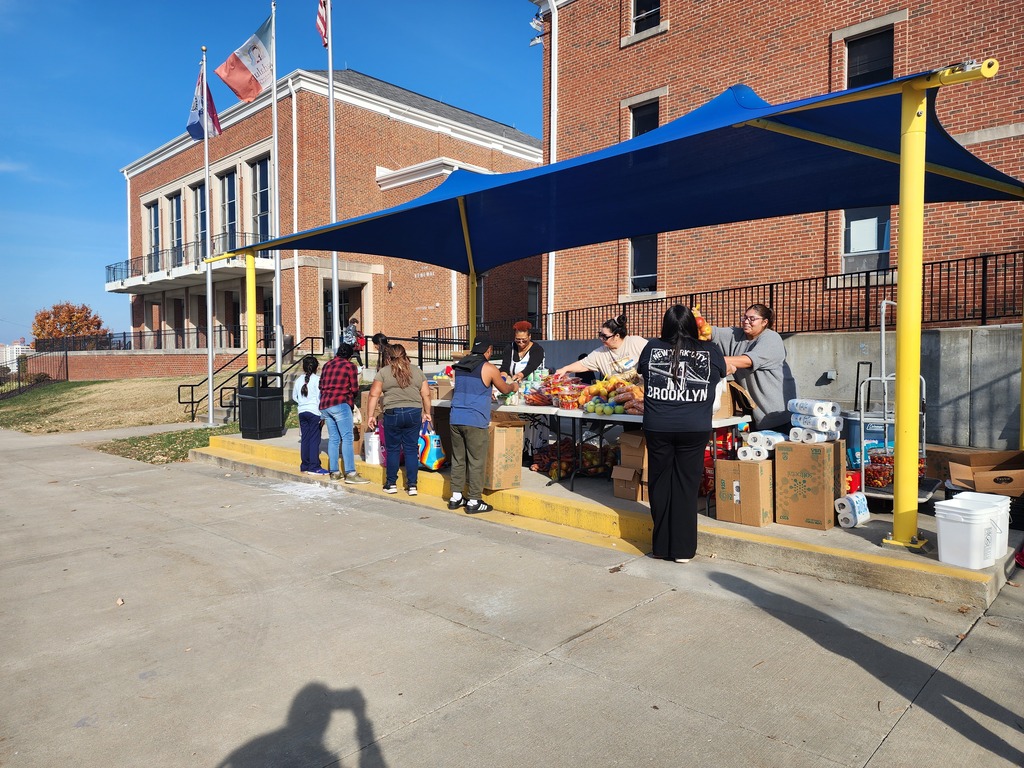 Volunteers are distributing food pantry items while people stand in line in an orderly manner to receive the products. The volunteers hand out food supplies as each person steps forward one by one, showing an organized and respectful food distribution process.