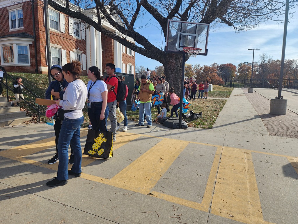 People are standing in line waiting to receive food assistance. A woman is recording their names on a list to help organize the distribution process. The scene shows an orderly food pantry line with volunteers managing registration.