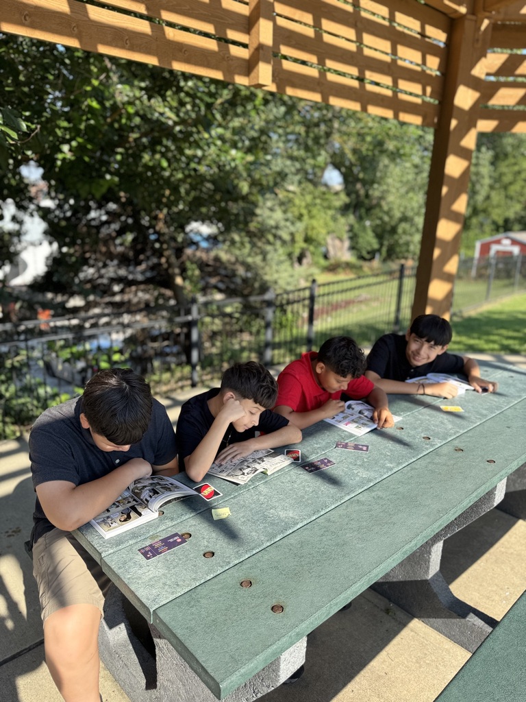 four students sitting at the  green picnic table reading their books, while the last student looks at the camera and smiles