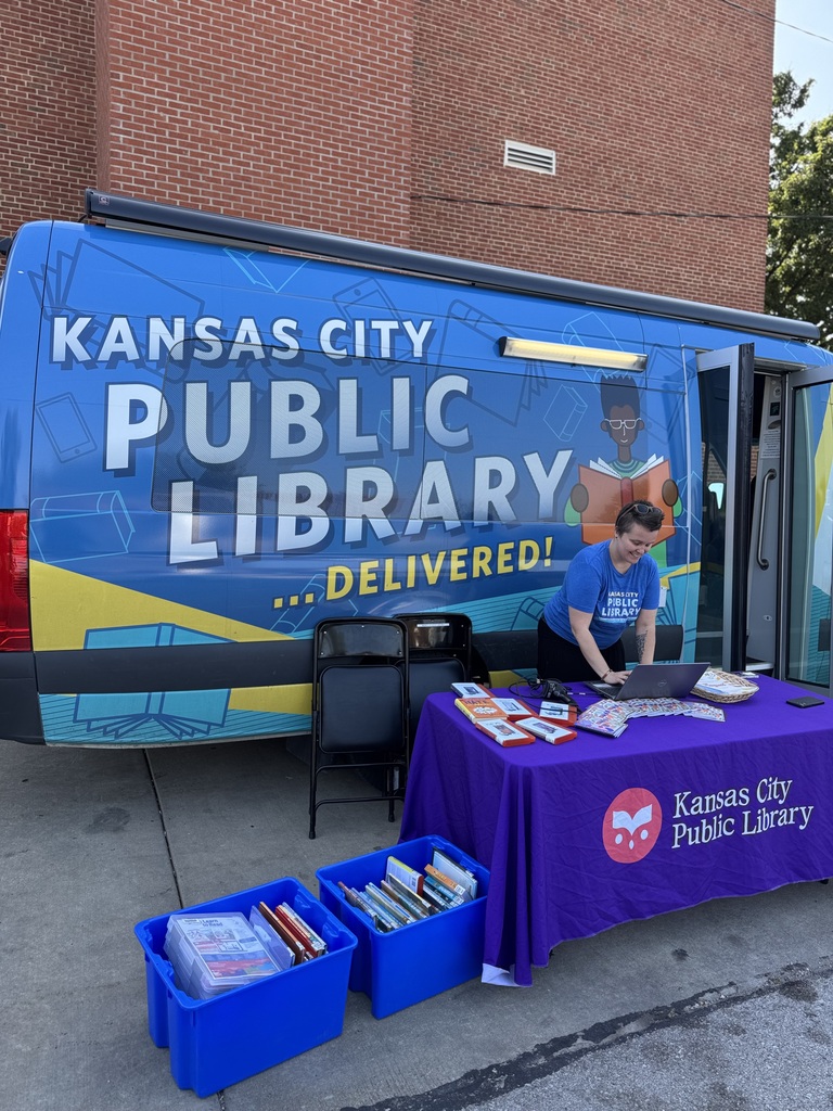 Blue van from the Kansas City Public library parked outside the school. There is a lady in a blue shirt organizing her table for students to check out books.