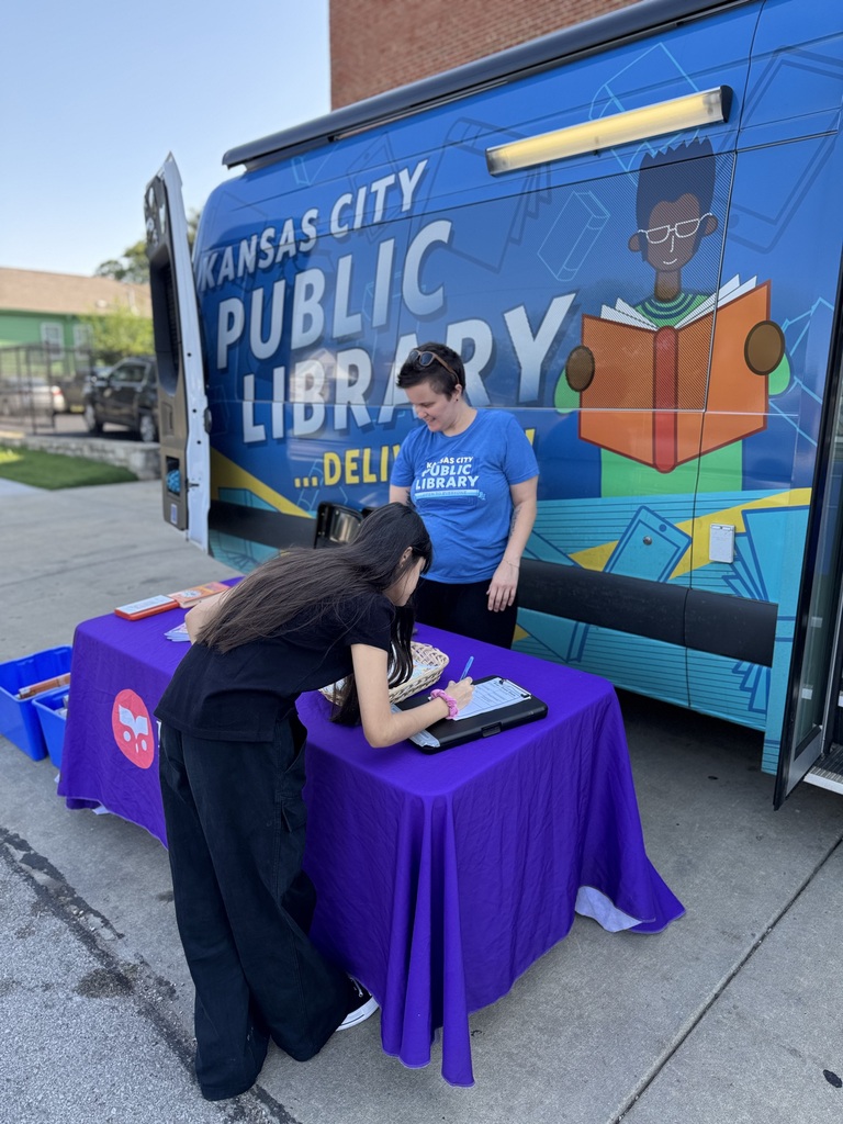 student wearing black uniform writing on a paper placed on the purple cloth table, blue library van parked outside the school 