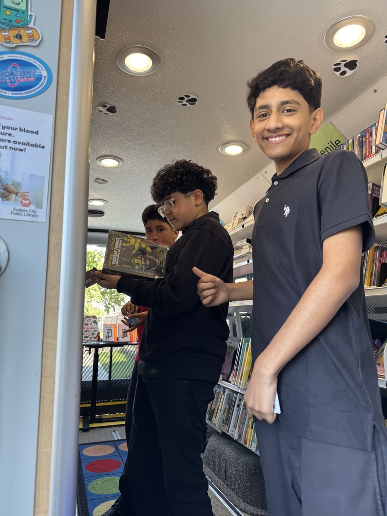 three students looking for books inside the bookmobile van. student closer to the camera is putting a thumbs up and smiling at the camera
