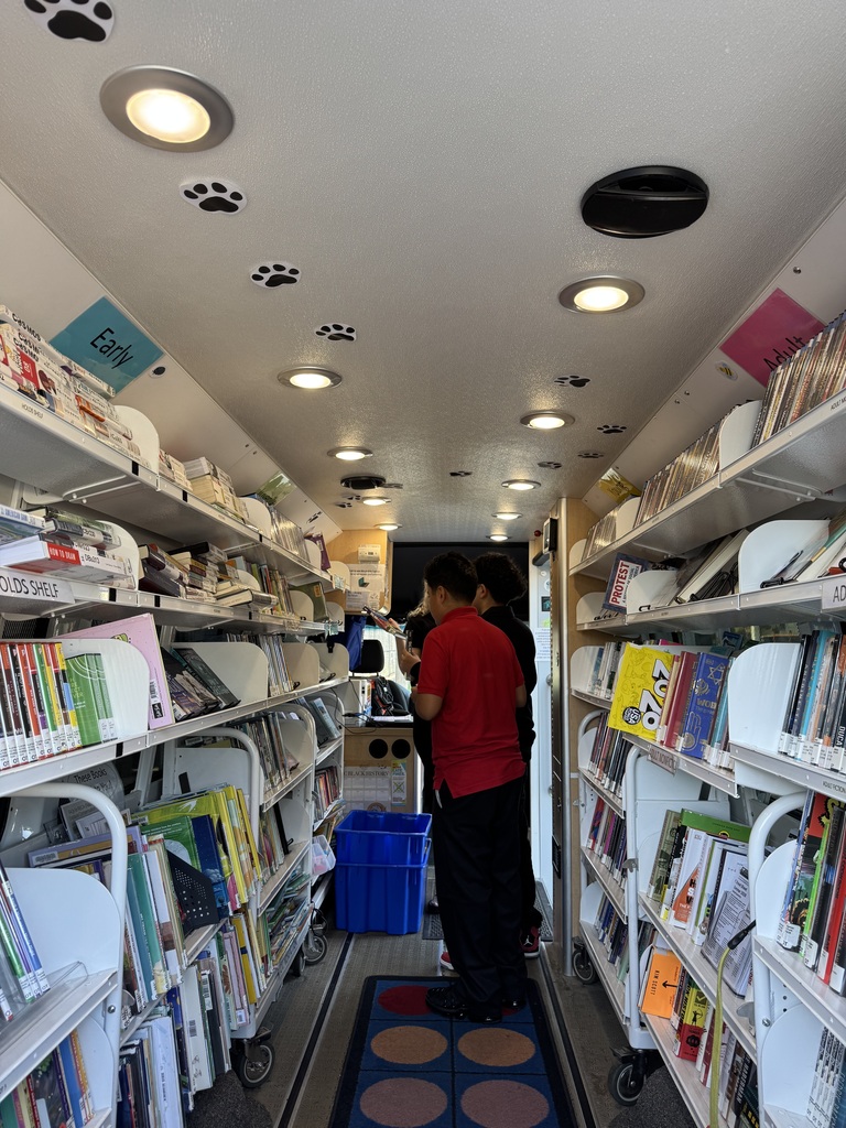 the interior of the bookmobile van, student in red polo shirt and black pants standing in line to check out a book