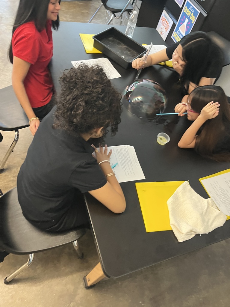 girls in black shirts and girl in red shirt are working together to make a big bubble on the black table 