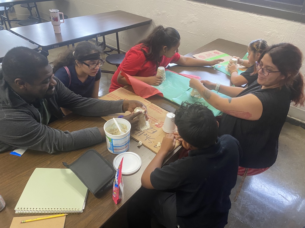 students working at a brown table in the cafeteria with teachers and doing a fun volcano experiment