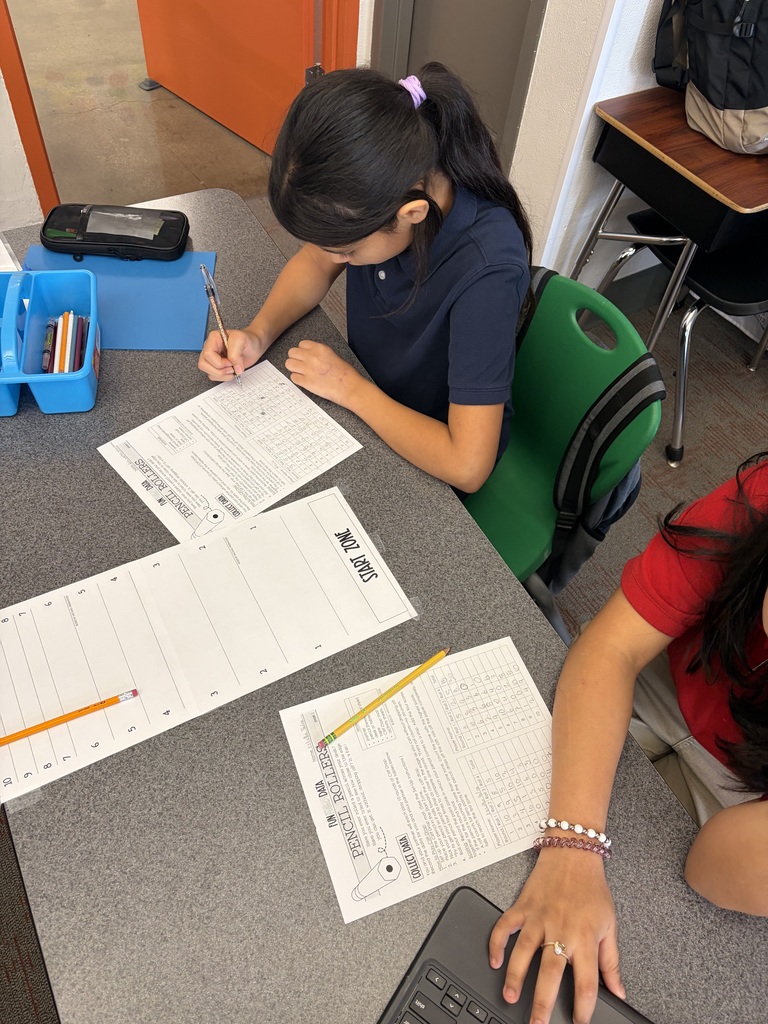student in navy blue is writing on her paper. another students hand is touching her computer.