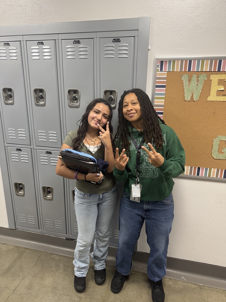 student wearing green and teacher wearing green, both with jeans on, posing in front of the lockers 