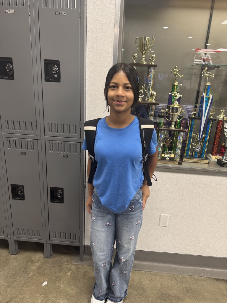student wearing blue shirt and jeans, wearing a black backpack, standing in front of the trophy box and lockers, posing for the camera
