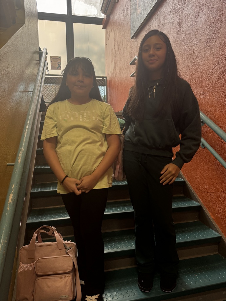 student in yellow shirt and black pants standing next to girl wearing all green, both posing on the green stairs and orange walls