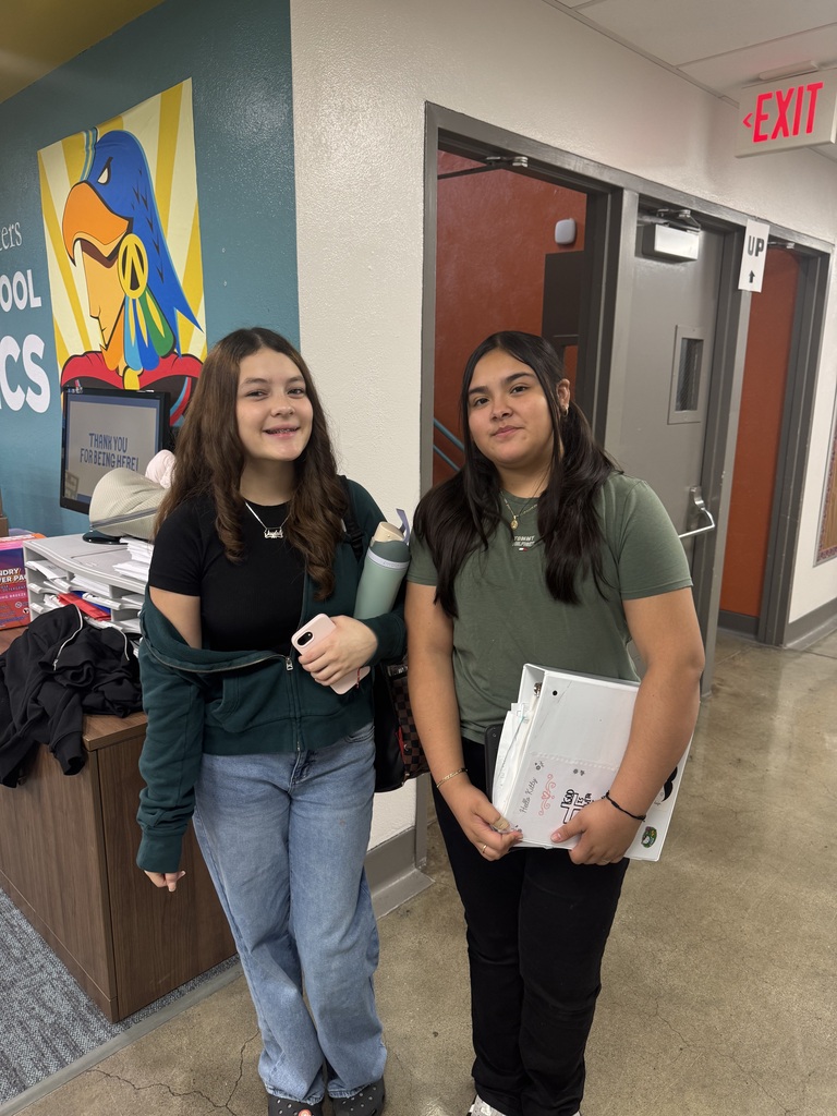 student in jeans, green sweater and black shirt, smiling next to her friend wearing a green shirt and black pants, standing in the middle of the hallway 