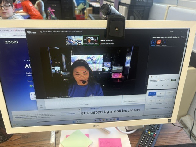 A computer screen, showing one of the members of the research team aboard the E/V Nautilus, speaking to the QUEST students