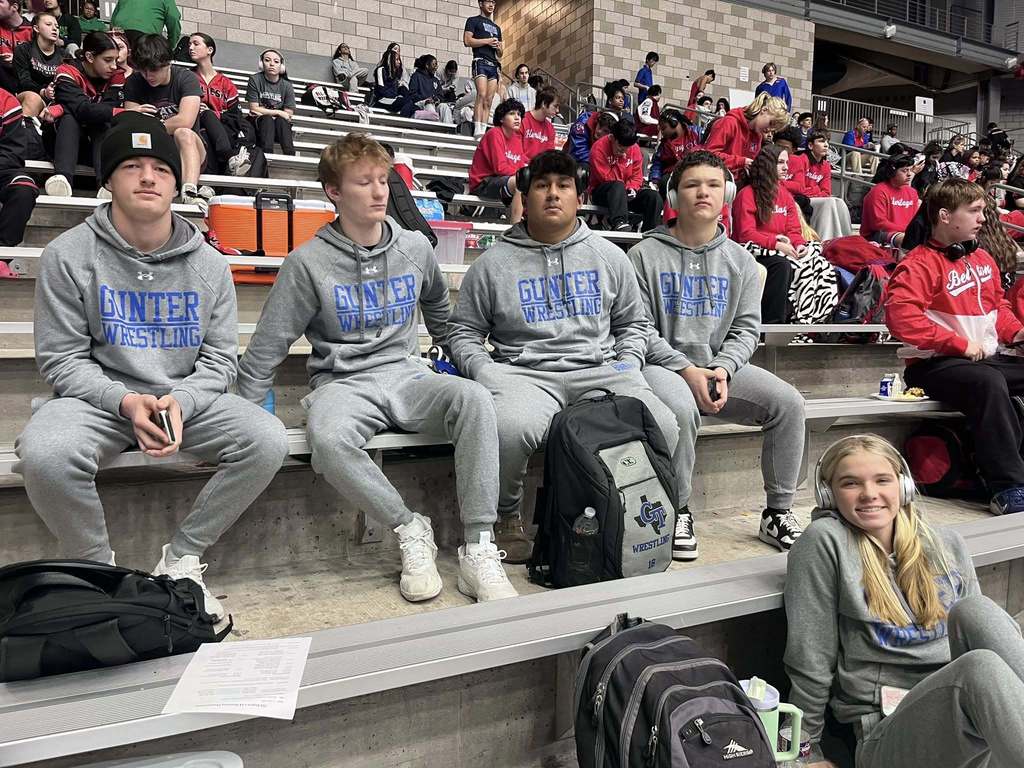 Five people wearing gray sweatshirts sit on bleachers, facing forward. One person holds a paper.