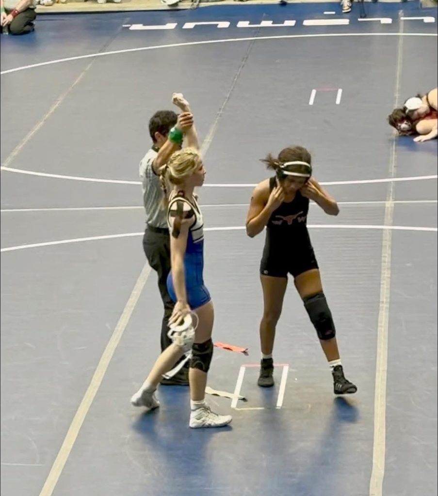 Two women in wrestling gear, one in blue, the other in black, stand facing each other. An official holds a raised green flag, signaling the match.