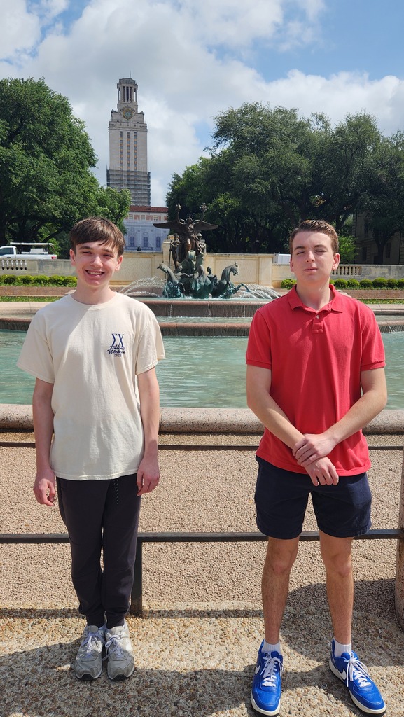 Two males pose for a photo with a fountain and greenery behind them.
