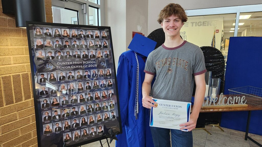 A young man stands in a room holding a certificate, with a graduation backdrop and a brick wall.