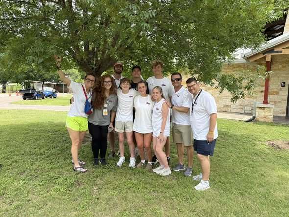 Group of people wearing white shirts and shorts, posing under a tree on a grassy area.